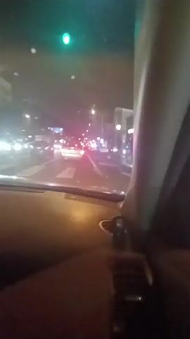The video shows a nighttime scene from inside a vehicle, likely a car, driving down a city street. The camera is positioned behind the dashboard, capturing the road ahead through the windshield. Streetlights illuminate the scene, casting light on the buildings and road markings. There are several cars visible in the distance, with their headlights creating streaks of light. At one point a police vehicle with flashing lights is visible. The street has a designated bike lane. The vehicle appears to be moving slowly or stopped in traffic. The camera angle shifts and moves slightly at several points. A building with a storefront is visible, with '#bellami' displayed on the outside.