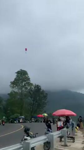 The video captures a mountainous landscape on a cloudy day with a paraglider floating in the sky. Below, along a winding road, are several parked motorcycles. There's a collection of small stalls set up with red and other colored umbrellas shading them. People are scattered around the stalls, some browsing, others standing and observing. Behind the road and the stalls, a steep, green mountain slopes up into the cloudy sky. The paraglider, visible against the grey sky, adds a sense of adventure to the otherwise tranquil scene.