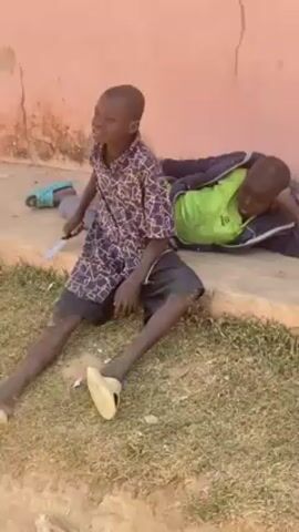 Two young boys are sitting by a wall. One is lying down while the other is sitting up. A third child walks over, carrying a bucket of water. They throw the water on the boy lying down.  The scene is an outdoor area with a wall and some grass.
