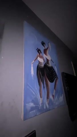 The video shows a painting hanging on a gray wall. The painting depicts two African women in traditional clothing, including white tops and black skirts with white stripes at the hem. They are holding white objects in their hands, possibly cloths or feathers. The background is a light blue color with white swirls. Next to the painting is a black object, possibly a TV or monitor, partially visible in the frame. The camera angle is looking up at the painting.
