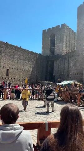 Here's a concise description of the video:

The video captures a staged knight fight at a castle. Two individuals, fully clad in medieval armor, face each other in a stone-paved courtyard surrounded by spectators behind a wooden fence. A man dressed in historical attire with a yellow shirt and straw hat stands to the side, presumably an announcer.  Behind the crowd, a massive stone wall and tower with multiple levels and viewing platforms loom. The sky is clear and blue, suggesting a sunny day.  A small yellow flag is visible near the announcer. In the distance, there are stalls set up, indicating a medieval fair or reenactment event. People stand on the castle walls and balconies to observe the scene below.