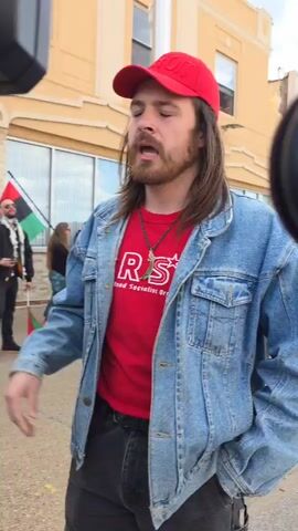 The video shows a man with long brown hair and a beard, wearing a red baseball cap, a red t-shirt with “Read Socialist Org” printed on it, and a denim jacket. He is standing outdoors on a street, and is speaking to someone off-camera. In the background, there are other people, a building, and a flag with red, black, green, and white stripes. The man appears to be at a protest or gathering.