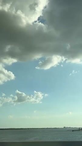 The camera faces upward at an expanse of sky. Below is an obscured landscape of land and water. The sky is a mix of clear blue and puffy white clouds, some larger and gray. The sun is shining and visible through the clouds, creating light flares. There is background music playing.