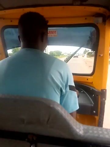 The video shows the interior of a yellow autorickshaw in Yelwa, Nigeria. The perspective is from the passenger seat, looking forward and slightly to the right. A man wearing a light blue tunic is driving the autorickshaw. His back is to the camera, and he is looking ahead through the windshield.

The scene outside the windshield appears to be a road in a rural or semi-urban area. There are trees and vegetation visible, as well as some buildings and possibly power lines in the distance. The road surface looks unpaved or in poor condition. The weather appears to be daytime, likely overcast or hazy, as the light is diffused.

A prominent detail inside the autorickshaw is a sticker on the driver's side window that reads "ARMY" with smaller text below it that is partially obscured but appears to say "UNIT OF MEN". The driver's hands are visible on the steering mechanism. The overall impression is a snapshot of everyday transportation in Nigeria. There are no strong indicators of emotion, but the focus is clearly on the act of driving and navigating the road.