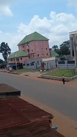 This video captures a street scene in Achalla, Nigeria, on a bright, cloudy day. The primary subject is a multi-story pink building with a green roof, which appears to be a commercial establishment or residential complex. In front of this building, there is a paved road with a few cars driving by.

To the left of the main building, there are smaller structures with corrugated metal roofs, possibly shops or market stalls. A tall, leafy tree stands to the left of the pink building, adding to the natural elements of the scene. On the right side of the road, there is a sidewalk and some fencing, with a few people visible walking. One person is walking on the sidewalk in front of the pink building, and another individual is seen further down the road.

The sky is filled with white, fluffy clouds against a light blue background, suggesting it is daytime, likely late morning or early afternoon. The overall impression is that of a typical day in a town, with everyday activities taking place. The road appears to be a main thoroughfare, connecting different parts of the community. There is no discernible text visible in the video.