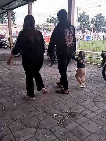 The video captures two adults and a young child walking together in a covered outdoor area. The ground is paved with interlocking bricks. The adults, dressed in dark long-sleeved tops and pants, walk side-by-side. The woman has long dark hair, while the man wears a backpack and flip-flops. A young child, wearing light-colored pants, a long-sleeved top, and a light-colored hijab, walks slightly behind the adults.

In the background, several motorcycles are parked, and other individuals can be seen. Beyond a fence, there is a green field dotted with colorful tents and decorations, indicating a possible public event or gathering. The lighting suggests it is daytime, possibly overcast or late afternoon. The scene is located in Matesih, Indonesia.