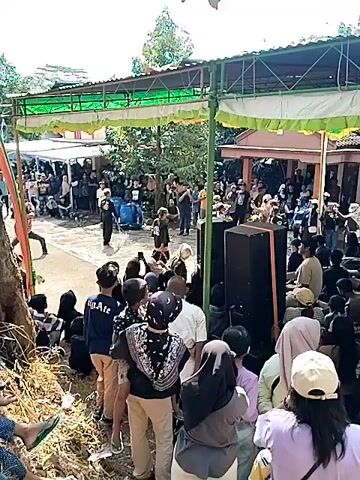 A crowd of people are gathered outdoors under a canopy. In the foreground, a large group of people, many wearing headscarves, are seated or standing, facing towards a stage area. The stage is set up with large speakers. In the mid-ground, a smaller group of people are on the stage, some appearing to be performing or speaking. The background shows more people and buildings, suggesting a community event or gathering. The lighting indicates it is daytime, with sunlight filtering through trees. The overall atmosphere appears to be one of a public event or celebration.