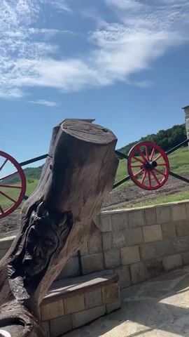 The video captures a sunny outdoor scene, panning from left to right across a rustic landscape. It begins with a close-up of a large, carved wooden sculpture or bench. In the mid-ground, a low stone wall serves as a fence, adorned with decorative red wagon wheels attached to black metal bars. Beyond the fence, green, tree-covered hills stretch under a bright blue sky with scattered white clouds. A white pickup truck is briefly visible parked behind the fence.

As the camera pans further right, a stone well-like structure with a circular base comes into view. This well has a wooden roof supported by sturdy wooden beams. Several colorful decorative plates hang from the underside of the roof; one prominently features a smiling yellow sun, while others display intricate patterns. On the stone surface of the well, a large clay jug is placed, bearing the text "БАЛКАНСКА БУРЯ" (Balkanska Burya, meaning Balkan Storm). Surrounding the jug are smaller decorative ceramic bowls or cups, arranged in an appealing display. The ground in the foreground is paved with irregularly shaped stone tiles, forming a patio area. The scene is devoid of people and suggests a tranquil, traditionally themed setting, characteristic of Medovo, Bulgaria.