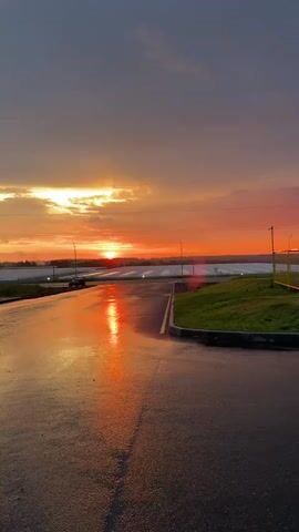 A vertical shot captures a sunset over a wet, paved surface. The sky is filled with hues of orange, yellow, and grey clouds. The sun, a bright orb, is low on the horizon, casting a vibrant orange reflection on the wet pavement. The wet surface appears to be a road or parking lot, with faint lane markings visible. To the left, a dark vehicle is parked. In the mid-ground, a line of white structures, possibly greenhouses, stretches across the landscape, bordered by a treeline. The right side of the frame shows a grassy area with a curb. The overall scene is tranquil and visually striking due to the dramatic colors of the sunset and its reflection.