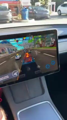 The video shows a car racing game being played on the center console screen of a Tesla. The perspective is from inside the vehicle.  Multiple Tesla cars are seen through the front windshield, parked near charging stations.  The game display indicates the player is in 6th place out of 6 racers, and is on the first of two laps. The game is colorful with a cartoonish art style. The player is actively steering their virtual vehicle, with steering controls displayed on the screen. Sounds from the game can be heard.