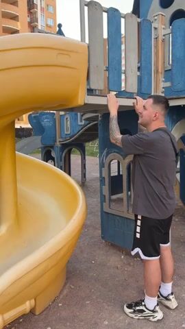 A man in a gray shirt and black shorts with white sneakers is standing facing a playground structure. He has a tattoo on his right arm and is wearing a black bag across his body. He is looking up and reaching his arms up to the playground structure. In the background, another man in a plaid shirt and dark pants walks by. The playground has a yellow slide and blue and white wooden walls. An apartment building is visible in the background.