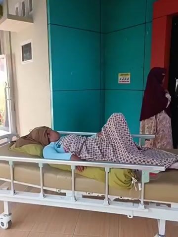 A woman lies on a hospital gurney, covered by a patterned blanket. She is wearing a light blue shirt and a headscarf. Her left leg is bent. A green bag is tied to the gurney near her feet. A person wearing a maroon headscarf stands to the right of the gurney, looking towards a doorway. A man in a white uniform and a face mask stands near the doorway, facing the woman on the gurney. Another woman in a yellow headscarf appears at the edge of the frame, to the left of the person in the maroon headscarf. The doorway has the number "3" above it. The environment appears to be a hallway or waiting area in a building, with teal-colored walls.