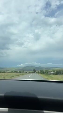 The video shows a view from inside a vehicle looking out through the windshield. The vehicle is on a paved road that extends into the distance. The road has white lines marking the lanes. On either side of the road are fields of grass and scattered trees. In the far distance, there are rolling hills and mountains under a cloudy sky. The clouds are gray and appear to be moving. There are a few small structures that appear to be buildings visible along the roadside in the distance.