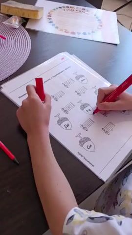 The video shows a young person sitting at a dark wooden table, engaged in a musical activity. The person is holding two red markers and appears to be working on a worksheet. The worksheet features musical notes and acorns, with some of the acorns labeled with Russian solfege syllables: "До" (Do), "Ре" (Re), and "Си" (Si). The person is coloring or marking some of the musical notations.

In the background, a digital piano is visible, suggesting a music lesson or practice session is taking place. To the left of the piano, a shelf with music sheets is partially visible. On the table, there's also a circular placemat, a yellow sponge eraser, and a pink object that might be clay or playdough. Another document with colorful illustrations and text is placed further back on the table.

The lighting suggests it might be daytime, with light coming from the right, possibly from a window, illuminating the scene. The atmosphere appears focused and educational. The overall setting is an indoor environment, likely a home or a music studio, given the presence of a piano and educational materials. The visible text on the worksheet is in Russian, and the bottom of the sheet has the text "Музыкальная". The document in the background has "Праздник ноты" (Holiday of the note) printed on it.