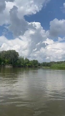 The video shows a wide, calm river flowing through a lush green landscape under a cloudy blue sky. Trees line both banks of the river. The water is a muddy brown color and reflects the sky and surrounding vegetation. The clouds are large and fluffy, with some appearing to be storm clouds. The overall impression is one of natural beauty and tranquility.