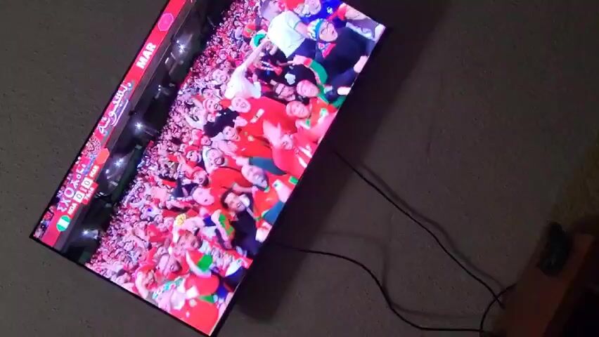 The video shows a TV screen mounted on a wall displaying a soccer game between Nigeria and Morocco, with the score 0-0. First, there is a shot of a crowd of fans in red and green attire. Then, the screen shifts to show two men embracing, one in a red soccer uniform and the other in a blue jacket. They are facing each other and tightly holding one another. There's a desk and a figurine in the background.