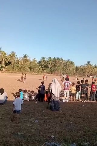 A group of people, mostly children and young adults, are gathered on a dry, dusty field. Some are playing soccer on the field, while others are watching from the sidelines. The spectators are standing and sitting in a loose group. The environment appears to be rural, with palm trees and vegetation visible in the background. The sky is clear and blue, suggesting it is daytime and fair weather. The scene is lit by natural sunlight.