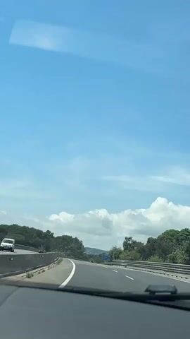 The video shows a driver's perspective while driving on a highway during daytime. The sky is blue with scattered clouds. The road is asphalt with white lane markings, and there are green trees and bushes on either side. A blue road sign indicates "C-31 Calonge Platja d'Aro 4". Cars and a truck can be seen driving on the highway.