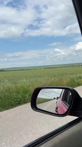 The video captures a serene view from a moving vehicle, looking out the passenger side window. In the foreground, parts of the car's interior are visible, including a dark gray window frame and a glimpse of a red car door in the upper right.

The dominant feature of the landscape outside the window is a vast expanse of green fields, likely crops such as wheat or barley, stretching towards the horizon. A narrow, grey asphalt road runs alongside the field, suggesting a rural or country setting. The sky above is bright blue with scattered white cumulus clouds, indicating a clear and sunny day.

Prominently featured in the lower center of the frame is the car's black side mirror. The mirror reflects the road behind the vehicle, showing the same grey asphalt receding into the distance, flanked by more green fields and a few distant trees under the cloudy sky. A portion of the red exterior of the car itself is also visible in the mirror's reflection.

The scene depicts a peaceful drive through the agricultural countryside near Sirakovo, Bulgaria. The bright lighting suggests it is midday or early afternoon. No people or discernible text are visible in the frame.