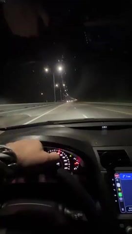 The video shows a first-person view from inside a car driving on a highway at night. The driver's hands are visible on the steering wheel. The dashboard lights and the car's navigation system are illuminated. Streetlights line the highway, and other vehicles can be seen in the distance. The road appears to be wet, and there are visible specks on the windshield, possibly from rain or insects.