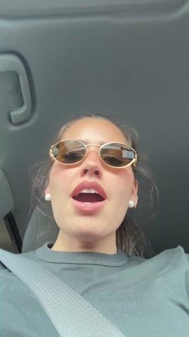A young woman is in a car, presumably driving, singing along to music. She's wearing a black t-shirt, has her seatbelt on, and is wearing gold-rimmed sunglasses and pearl earrings. The car's interior is visible, including the ceiling and a light fixture. The view is from a slightly low angle, looking up at the woman.