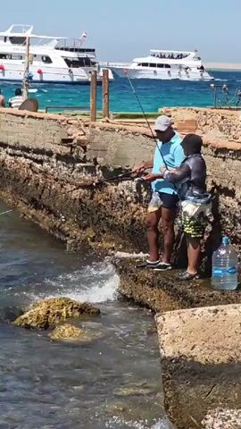 Two people are fishing off a rocky pier. The person on the left is an adult male wearing a blue t-shirt and black shorts. The person on the right is a child wearing a dark gray t-shirt and patterned shorts. Both are holding fishing rods. In the background, there are two white boats on the water. The water is blue and there are waves crashing against the pier. There is a large clear plastic bottle of water on the pier to the right of the child. The sky is clear and blue.