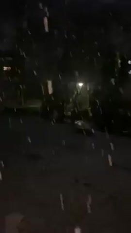 The video depicts a snowy night scene. The camera is stationary, showing a view from what appears to be indoors, looking out at a dark street. Large, thick snowflakes are falling heavily, obscuring the background. In the distance, a street light provides a faint glow, illuminating a stretch of road. There are dark shapes that resemble trees and buildings faintly visible behind the falling snow. The overall impression is one of a heavy snowfall at night.