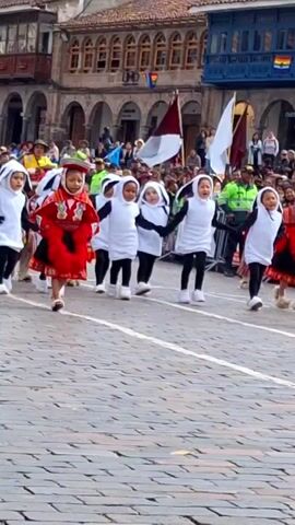 Children in costumes are marching in a parade on a cobblestone street. The children in the foreground are dressed as penguins, with white bodies and black wings. One child in the center is wearing a red and white traditional outfit. There are adults in the background watching the parade, and buildings with balconies line the street. Flags are visible, including one with a rainbow design.