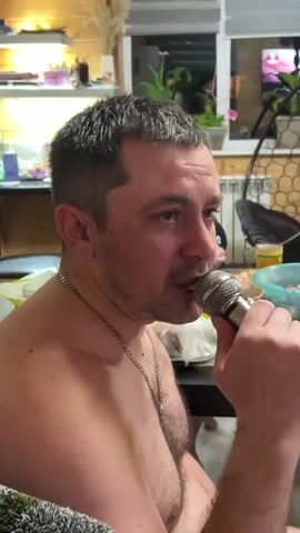 A medium shot captures a fair-skinned man, shirtless and wearing a thin silver chain necklace, holding a microphone to his mouth and singing. He has short, salt-and-pepper hair and is looking slightly off-camera to his right. The backdrop reveals a domestic interior with a shelf displaying various items, a window, and a wicker chair. In the distance, a television screen is visible. The scene is warmly lit, suggesting indoor lighting.