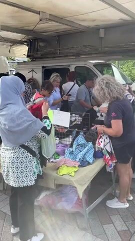 A street market stall displays an assortment of clothing items, predominantly bras and underwear, neatly folded on tables. Several women are browsing the merchandise. One woman, wearing a light blue hijab and a patterned blouse, examines a red garment. Another woman with curly red hair and a patterned shirt is also looking at the clothing. A man stands behind a table on the right, facing away from the camera. In the background, a white van is partially visible. The market appears to be set up outdoors under a white awning, with a brick-patterned walkway in the foreground. The overall atmosphere suggests a casual shopping environment.