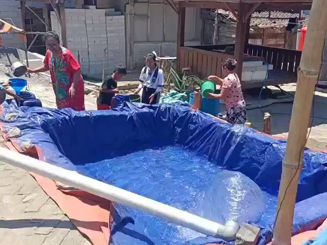 A group of people, including adults and children, are filling a large blue tarp with water. The tarp is spread out on the ground and appears to be forming a makeshift swimming pool. Some individuals are pouring water into the tarp from buckets, while others are observing. The environment seems to be outdoors, possibly in a residential or construction area, with buildings and building materials visible in the background. The scene suggests a communal effort to create a place for recreation.