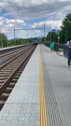 The video begins with a view of a train approaching a station platform. Two people stand on the platform, facing the approaching train. One person, wearing a bright green jacket, is looking towards the train. The other person, partially obscured, is also looking in the same direction. The train, a blue and white locomotive, is on the tracks to the left of the platform. The platform is paved with gray tiles and has a yellow tactile warning strip along the edge. In the background, there are more train tracks, overhead electrical lines, a blue sign labeled "2A", trees, and hills under a cloudy sky. The camera angle is low, looking slightly up towards the train and the sky. The train is moving towards the camera and appears to be slowing down as it approaches the platform.