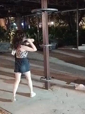 A woman in a diamond-patterned top and striped shorts hits a metal pole with a hammer under a metal roofed structure. Another person stands near the woman, partially visible. The area appears to be under construction, with loose construction debris and material scattered around. The scene is lit artificially, likely at night.