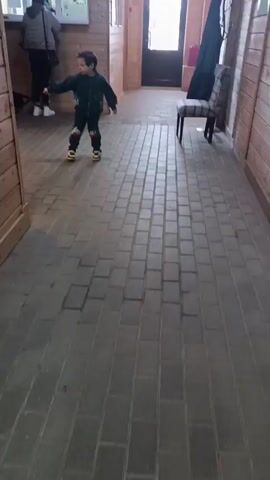 A young child is walking through a building carrying a black riding helmet on a short whip-like riding crop. The child is wearing a green and blue tracksuit and appears to be holding the whip loosely. Behind the child is a person standing at a ticket window, appearing to purchase something. The floor is paved with rectangular gray stones, and the walls are paneled with wood. In the background, there is a partially visible door. There is a wooden door to the left of the child. There is a chair to the right of the doorway in the background.