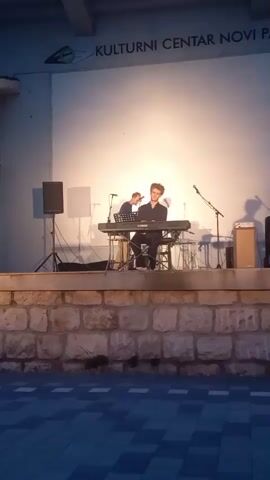 The video shows a young man playing a keyboard on an outdoor stage. He is seated on a stool behind a Yamaha keyboard, with sheet music displayed on a stand. He is dressed in dark clothing and appears focused on his playing. The stage is elevated on a stone wall, with speakers and microphones set up for the performance. Behind the stage is a large white wall with text that reads "KULTURNI CENTAR NOVI PA". The lighting suggests it is evening or dusk, with the stage area illuminated by spotlights. The overall scene conveys a sense of a musical performance taking place at a cultural center in Novi Pazar, Serbia.