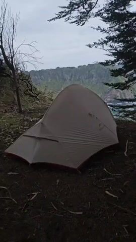 The video shows a tent situated in a camping spot overlooking a cove. The tent is grey and red, and appears to be a two-person tent. It is nestled among trees and roots on a dirt surface. The camera pans to show the cove, which features a sandy beach and clear teal water with gentle waves. Lush green vegetation covers the surrounding cliffs. The sky is overcast and grey, suggesting a cloudy day.