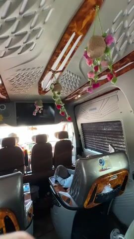 This media file captures the interior of a customized van, likely a passenger van due to the multiple rows of plush, brown leather seats. The van appears to be stationary. The scene is lit by natural light filtering in from the front windshield, suggesting it is daytime. The ceiling of the van is elaborately decorated with a textured pattern and ornate wooden trim. Hanging from the ceiling are two decorative items: one is a woven ball with pink and green crocheted flowers, and the other is a cluster of pink crocheted flowers with green stems.

In the foreground, two passenger seats are visible, upholstered in grey and accented with wood-like trim. A person is reclined in one of these seats, covered with a light blue blanket or garment, suggesting a passenger is resting. A flat-screen television is mounted on the wall behind the driver's section.

Further back, three more rows of passenger seats are visible. The front windshield offers a glimpse of the outside, showing the top of what appears to be another vehicle, possibly indicating the van is parked in a lot or on a road. The overall impression is one of a comfortable, perhaps luxury, personal transport vehicle. The decorative elements, like the crocheted flowers and the wood trim, add a unique and personalized touch to the interior. No specific weather conditions are discernible, but the ample natural light suggests it is not heavily overcast. There is no discernible text in the media file. The setting strongly suggests a customized interior for long-distance travel or a mobile living/working space.