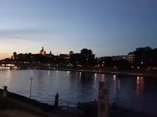 This is a video of a river at dusk. The sky is a deep blue with some light clouds. The water reflects the lights from the buildings on the opposite bank, which include a large, ornate building with towers and illuminated windows. There are trees and other buildings visible along the shoreline. In the foreground, there is a walkway or pier with some railings and a sign. A person is visible standing on the edge of the walkway. The scene appears to be in a city.