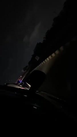 The video shows a dark interior view from a car, presumably at night. The rearview mirror is visible in the center of the frame. Outside the windshield, there are vague shapes suggesting buildings or trees silhouetted against a dark sky with some visible clouds. The lights of other vehicles are visible in the distance, adding some illumination. The overall scene is dark, with limited visibility. The camera is still and there is no discernible human activity within the car.