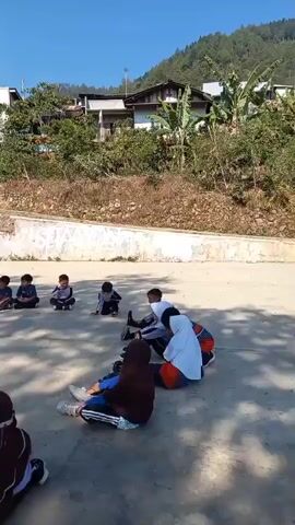 A group of young children are sitting and lying on a concrete ground outdoors, some in school uniforms. The background shows a hillside with houses and trees, under a clear blue sky. One child in a white hijab is lying on their back, while another in a brown hijab sits facing away from the camera. Several other children are seated in a semicircle, some looking towards the center. The scene appears to be a schoolyard or open recreational area.