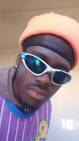 A close-up, low-angle shot features a young Black man wearing a peach-colored beanie over a black du-rag. He sports futuristic silver-rimmed sunglasses that reflect his surroundings and a purple-striped jersey with a Mamelodi Sundowns emblem on the left breast. He raises both hands to display his middle fingers. The camera is slightly shaky. The background is plain, showing a beige-colored wall and ceiling. Music plays in the background. The man then lowers his middle fingers and lowers the sunglasses slightly down his nose.