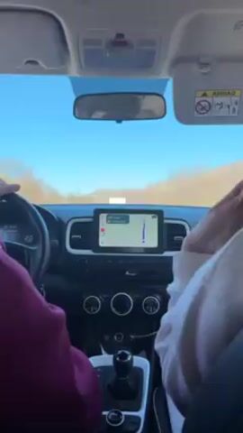 The video shows the interior of a car driving on a road. The driver, wearing a maroon sweatshirt, is seen behind the steering wheel. The passenger, wearing a white sweatshirt, is filming with their phone. The car's navigation system is visible on the dashboard, displaying a route. The road ahead is clear under a bright blue sky. Trees are seen on the sides of the road.