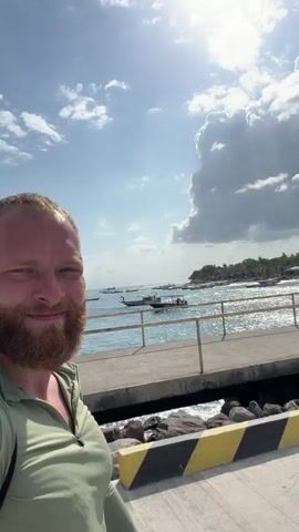 A man with a beard films himself with a selfie stick on a pier. Behind him, several people are walking away from the camera. The pier has a concrete walkway with yellow and black striped barriers on the left side. The water is visible to the right, with several boats docked. In the distance, there are buildings and palm trees. The sky is partly cloudy with sunlight. The man smiles and waves to the camera.