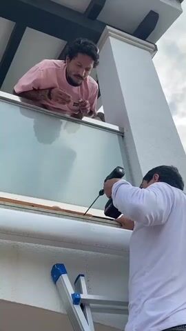 A man in a pink t-shirt leans over a balcony railing, looking down at another man who is using a cordless drill to bore a hole in a wooden frame. The second man is standing on a ladder and wearing a white long-sleeved shirt. The drill is pointed at the frame, and sawdust is visible. The scene appears to be outdoors, with a cloudy sky in the background.