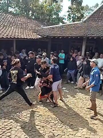 The video shows a crowd gathered outdoors, likely in a village, watching a performance. The ground is paved with rough, uneven stones. Several people are dressed in traditional or costume attire, with one individual in the foreground wearing a black shirt with a design and dark pants, performing some sort of dance or martial art. Another person, dressed in blue, stands watching. In the background, more people are visible, some seated and some standing, observing the performance. A building with a tiled roof and a porch is behind the spectators. The atmosphere appears festive, with flags also visible. The scene is lit by daylight.