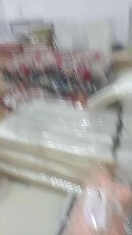 The video shows a dimly lit room with a desk covered in various items. On the desk, there are stacks of fabric, some of which appear to be covered in plastic. There are also books, papers, and a printer. A calendar hangs on the wall behind the desk, along with a framed picture. The room has white walls and appears to be an office or storage area. The lighting is dim, creating shadows and making it difficult to see some of the details clearly. The camera is handheld and the footage is slightly shaky. The video pans across the scene, showing different parts of the desk and the surrounding area.