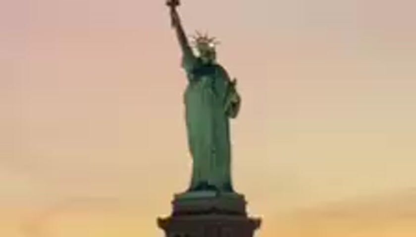 The video displays the Statue of Liberty at dusk. The sky is a gradient of soft pink and orange hues, indicating the sun has recently set. The statue, a copper-green icon, stands tall against this backdrop, its torch illuminated. The camera is positioned from a distance, likely across the water, offering a broad view of the monument. No people are visible, and the scene is static, focusing solely on the iconic landmark. The weather appears clear, with no visible clouds obscuring the sky. The setting is clearly Liberty Island, New York Harbor, with Jersey City implied as the vantage point. The overall mood is serene and majestic, highlighting the grandeur of the statue during twilight. There is no visible text within the video frames.