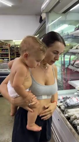 A woman is holding a baby and looking at shelves of food products in a store. The woman is wearing a grey sports bra and black shorts, and the baby is wearing a diaper. The shelves are filled with various packaged food items, some of which appear to be noodles or pasta. The woman is pointing at or holding one of the packages. The environment appears to be a grocery store or market with refrigerated displays.