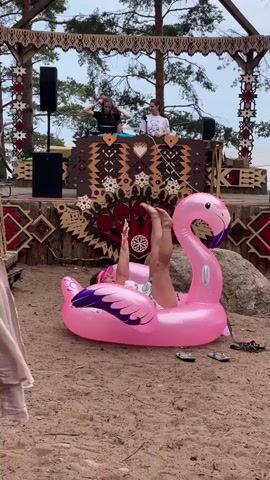 A video captures a lively outdoor scene with a stage adorned with geometric patterns. Three people are on the stage, two appear to be DJs with equipment set up. A person is seated on a large, inflatable pink flamingo on the sandy ground in front of the stage. The person on the flamingo is dressed in casual clothing, including denim shorts and a patterned top, and is wearing a pink visor. This person is playfully interacting with the camera, at one point raising an arm and smiling. The background consists of trees and a cloudy sky, suggesting an outdoor event, possibly a festival or party. The overall atmosphere is relaxed and festive.
