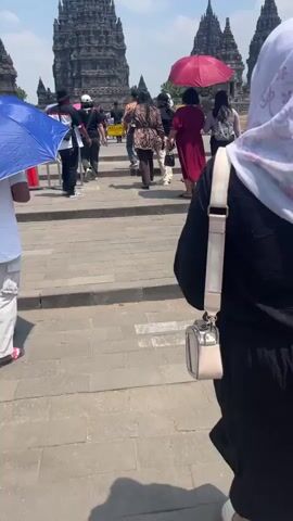 A group of people walk past ancient stone temples on a sunny day. In the foreground, a person wearing a white top, white pants, and sandals walks to the left, holding a blue umbrella. To their right, another person, whose face is not visible, wears a black top and a cream-colored purse with a fluffy keychain. Several individuals in the background are walking towards the temples, some holding umbrellas. One person in the middle has a pink umbrella, and another is wearing a red dress. The temples are tall and dark, with multiple spires. A yellow sign with a red circle and line through a cigarette is visible on the right. The ground is paved with stone.